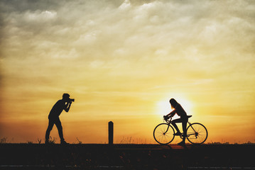 Silhouette of boyfriend taking pictures of girlfriend cyclists. couple are cycling happily in time for sunset.