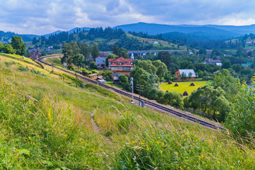 Naklejka premium A path on a slope overgrown with grass and flowers leading to the railway below which makes a smooth turn around the village with houses.