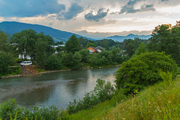 Fototapeta premium roofs of rural houses among green trees on the bank of the river with a white minibus parked next to it against the backdrop of distant peaks and a gray cloudy sky. place of rest and tourism