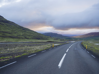 Asphalt road curve through empty northern landscape with green grass colorful hills, water puddles and sunset dramatic sky, way to the mountains in Iceland western highlands, copy space