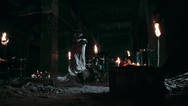 Spooky psycho in a mask and a white hoodie smashes a lantern with a baseball bat while a drummer plays drums in an abandoned factory with lighted torches on background