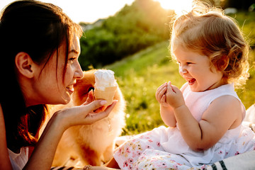 Mom with her 2 years old daughter sitting on the blanket and eating ice cream in the park. Sunset. Cute dog looking on them. Good relations of parent and child. Happy moments together.