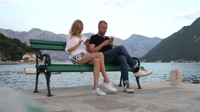 young couple sitting together in phones on a bench near a mountain lake