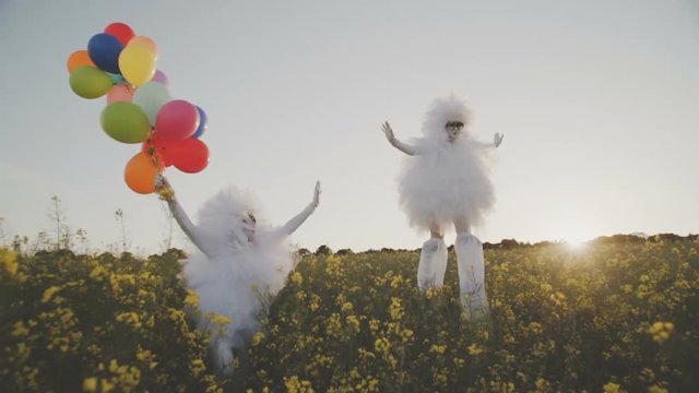 Two funny clowns mime in white air suits on stilts and with colored balloons walk around the field and have fun.