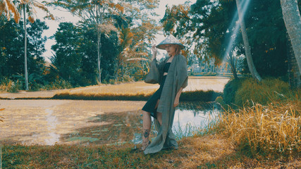 Beautiful fashion girl in kimono and traditional Balinese straw hat posing in tropical rice fields...