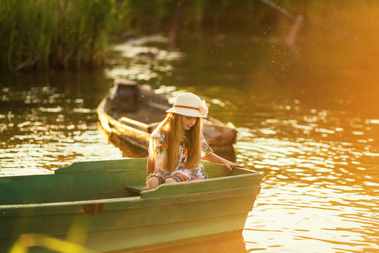 Cute little girl in a hat sitting in a boat on a lake at sunset