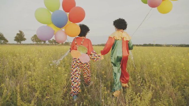Two funny clowns in colorful costumes with colored balloons are having fun in the field.
