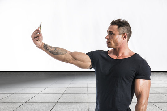 Handsome muscular young man taking selfie with cell phone while striking bodybuilding pose, isolated