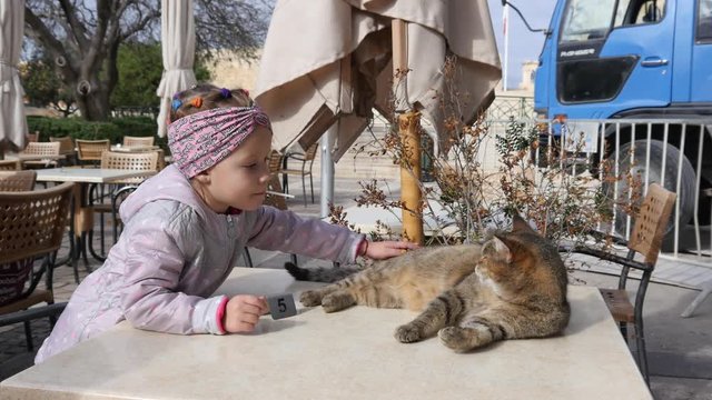 Valletta, Malta - Child girl play stroking a cat lying on street cafe table - kids and animals
