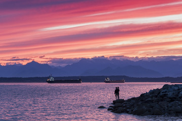 Fototapeta na wymiar Silhouetted couple at sunset looking out at a glorious sunset with cargo ships next to the Olympic Mountains on Puget Sound