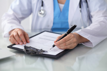Doctor woman filling up medical form while sitting at the table, close-up of hands