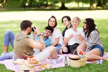 friendship, leisure and technology concept - group of happy smiling friends with non alcoholic drinks photographing at picnic in summer park