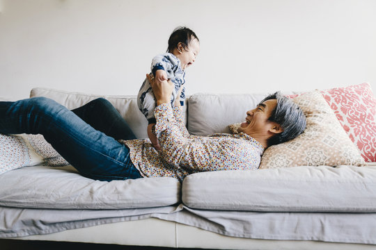 Happy Grandfather Playing With Grandson While Lying On Sofa At Home