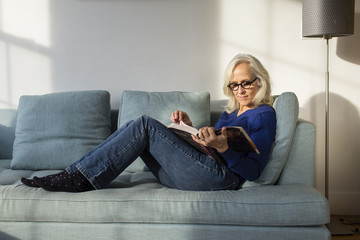 Full length of senior woman reading book while resting on sofa at home