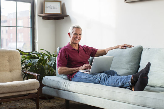 Full length portrait of mature man using laptop computer while resting on sofa in living room at home