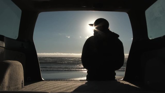 With the sun at his eye, model released man looks out at the Pacific Ocean from the back of his wagon parked on sandy beach in Oregon.