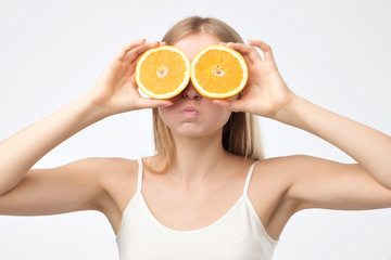 Beautiful young blonde woman in white shirt holding piece of orange near her eyes like glasses blowing her cheeks while standing against white background. Concept of eating fruits and healthy diet