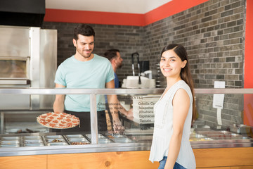 Happy customer standing near pizza counter with chef holding pizza in background