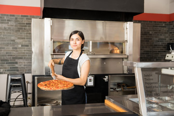 Young woman holding baked pizza on shovel and placing on counter