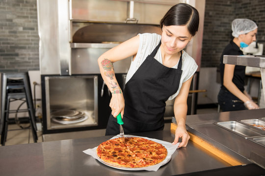 Young chef cutting pizza by using cutter in kitchen counter