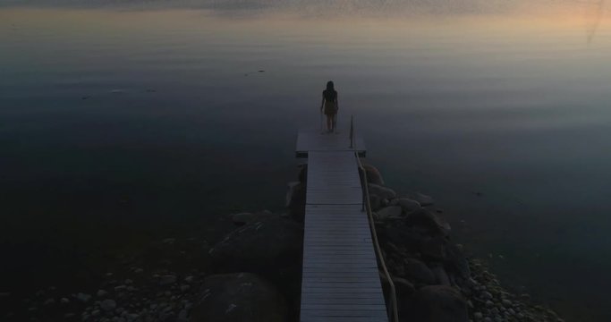 Woman on a wharf, C4K aerial descending tilt view a woman, standing at a wooden dock, enjoying the sun and a warm summer sunset, on a sunny evening dusk, in Hanko, Uusimaa, Finland