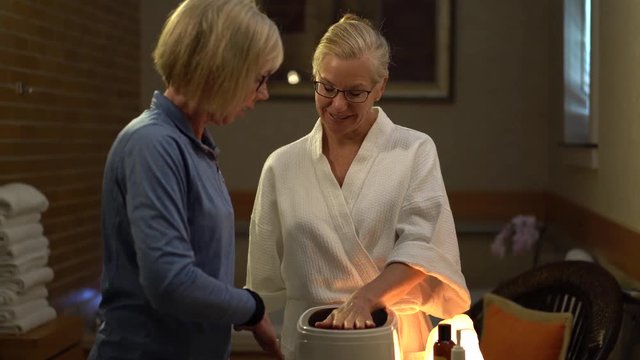 With attendant helping her, a mature woman dips her hand into hot paraffin wax for a skin treatment.