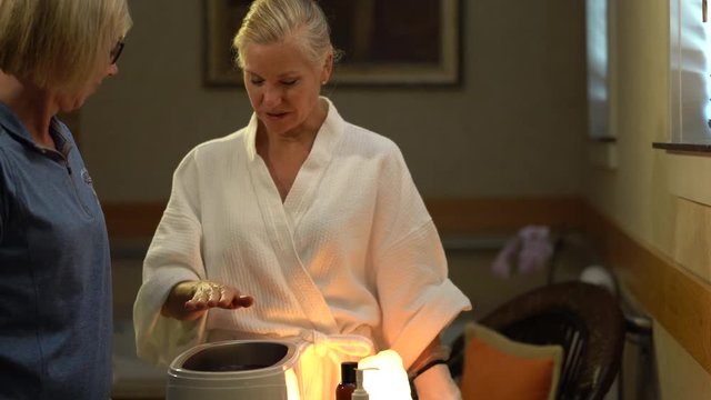 Pretty blonde woman smiles to the therapist and puts her hand into the hot paraffin for her hand skin treatment.
