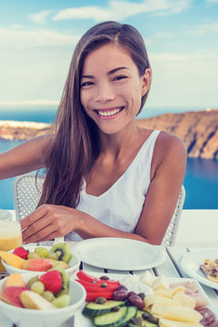 Woman eating healthy breakfast at restaurant table with Mediterranean sea view background from hotel terrace. Smiling Asian girl, vegetarian fruits and vegetables food retreat.