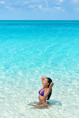 Bikini body beach swimwear model swimming in idyllic turquoise ocean water relaxing under the sun. Asian woman touching her healthy hair in swimsuit tanning body.