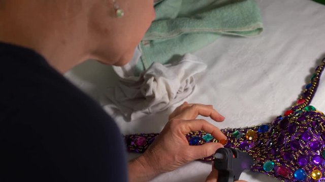 Looking down onto a decorated bra, a woman uses a glue gun to glue plastic rhinestones into place.