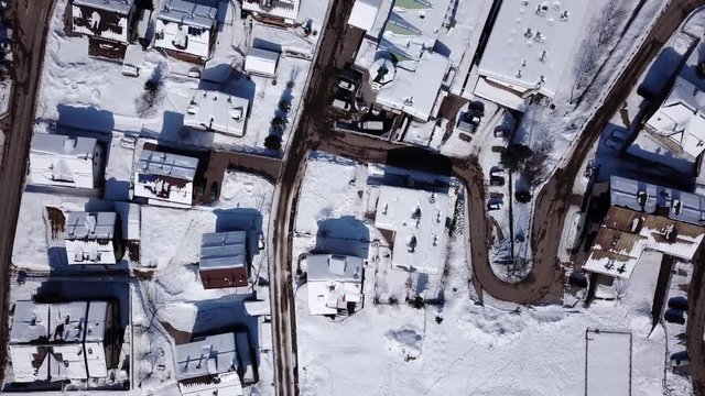 Topdown drone shot over a village in the alps in winter. Small houses and one bigger building. Snowy landscape and rooftops but dry roads.