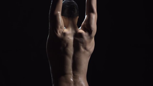 Studio shot with rear view of unrecognizable male athlete in swimming cap standing isolated on black background and stretching before practice