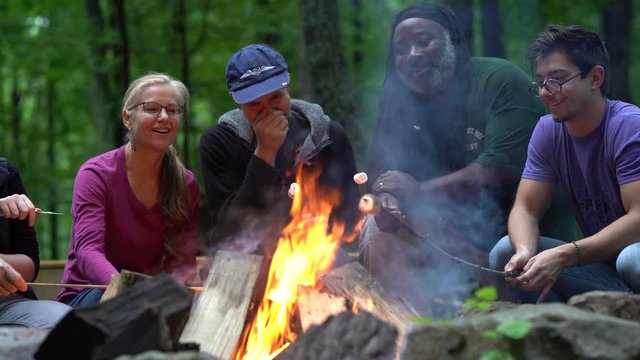 Family and friends sit around a campfire and roast marshmallows as camera slides to the left.