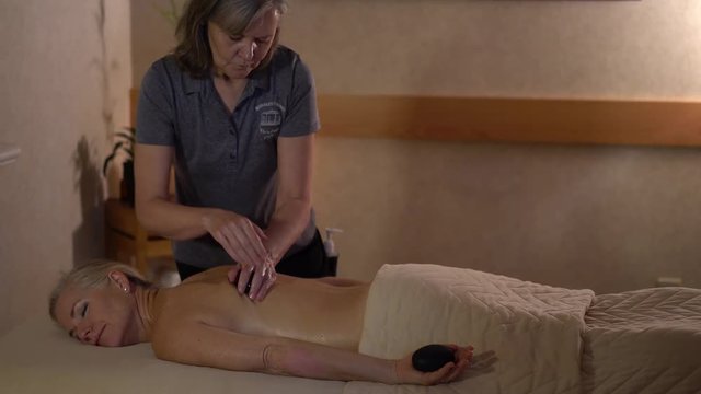 Massage therapist uses hot stones to rub the side of a blonde woman’s back.