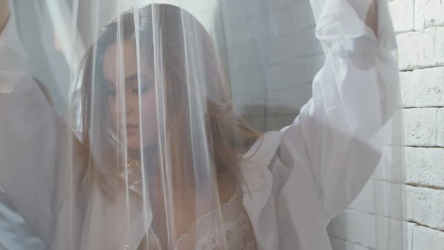 PAN of beautiful young woman in lingerie and shirt standing behind sheer curtain and moving seductively