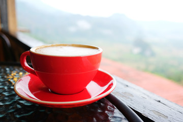 A red cup of coffee on the table with nature of mountain and flowers field background
