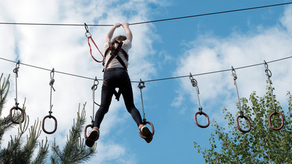 A girl in black jeans and a white T-shirt passes an obstacle course above the trees against the sky with clouds. Extreme entertainment. Summer rest.