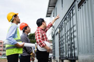 foreman and employee holding blueprint on hand and pointing finger to building. engineer checking construction process in construction site area..