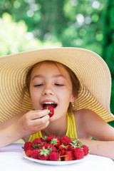 Portrait of Cute teen girl in big hat is eating strawberry at summer day