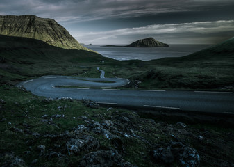 a curvy road down a green hill surrounded by grass fields heading to the ocean with an island in the center of the frame and a dramatic sky on the Faroe Islands during sunset