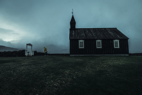 a black church with white window frames and a gate infront of the church taken from the side on a misty rainy windy dark moody day in Iceland with a lonely man in a yellow rain coat walking out 