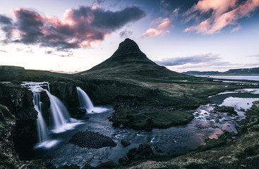 Fototapeta na wymiar Kirkjufell Mountain in Iceland in the background with three waterfalls in silky look in the front with a river flowing around a mountain into the ocean and a colorful pink and purple sky with clouds