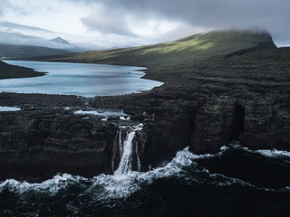 A lake above the rough ocean with waves smashing on high cliffs on a moody misty day with a dramatic sky and mountains in the background on the faroe islands