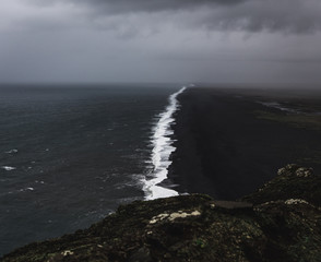 Bird perspective on a coastline in Iceland with a black sand beach with black and white contrasts between the waves and the beach while a storm is approaching with dark clouds in the sky