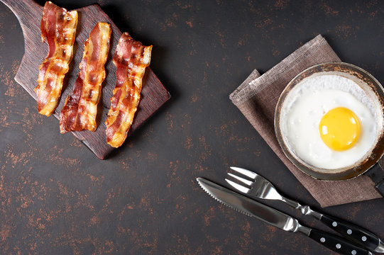 Fried bacon on cutting board and egg with fork and knife. Breakfast top view, isolated on black background.