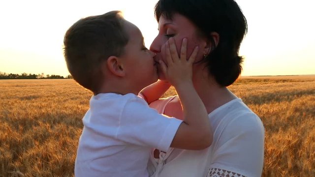 A happy mother holds a child in her arms in a field of wheat against a sunset background. The baby and mom are kissing. The concept of a happy family
