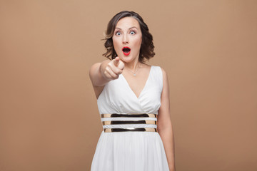 Fototapeta premium Wow! Shocked adult woman pointing finger at camera. Emotional expressing woman in white dress, red lips and dark curly hairstyle. Studio shot, indoor, isolated on beige or light brown background