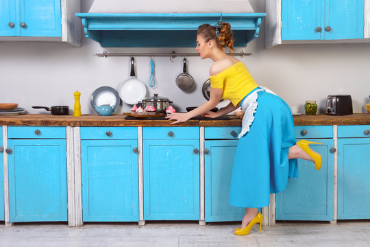 Retro pin up girl woman female housewife wearing colorful top, skirt and white apron and yellow high heels looking at cooked sweet cupcake standing in the kitchen with blue cabinets and utensils.