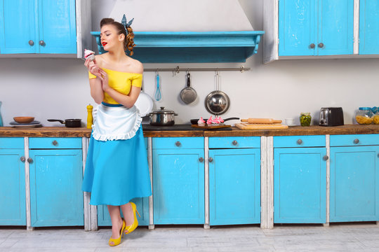 Retro pin up girl woman female housewife wearing colorful top, skirt and white apron and yellow high heels holding cooked sweet cupcake standing in the blue kitchen with blue cabinets and utensils.