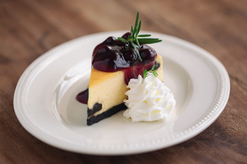 Delicious and sweet blueberry New York cheesecake on white plate served with whipped cream on wood table in side view with copy space. Homemade bakery for cafe and restaurant or birthday cake.